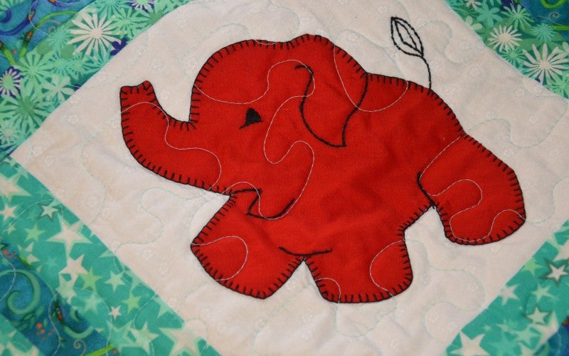 A white square on a teal quilt containing a quilted-on red elephant.