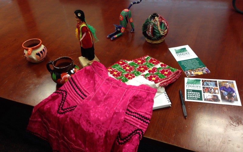 Traditional art pieces, including two mugs, a doll, a white embroidered shirt, a pink skirt, and a cat figure all sit on a table.
