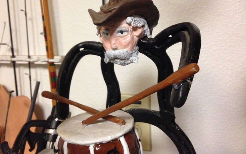 A metal art piece in which an older man wears and brown cowboy hat and plays the drums.