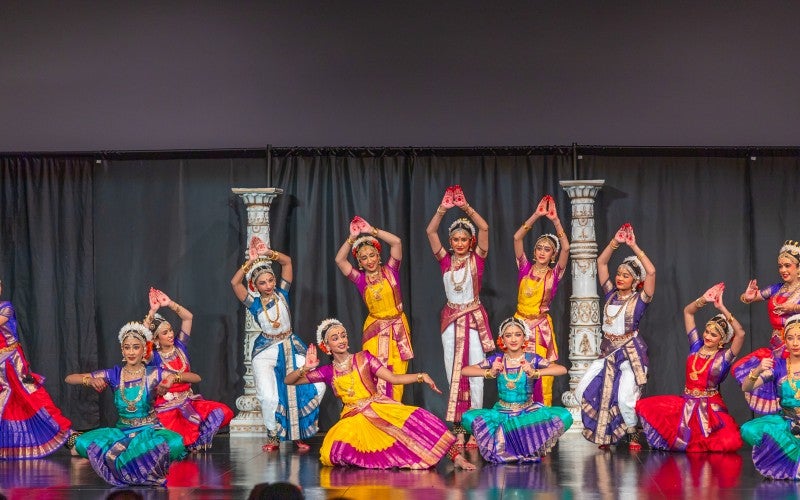 13 dancers are posing in expressive dance gesture while performing on stage. All of them in traditional Kuchipudi dance attire.