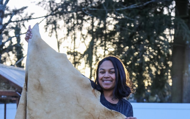 Eleeziaa stands holding a tanned hide, which is as tall as she is
