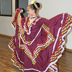 A girl dances ballet folklorico while wearing a traditional Mexican dress of maroon color and multicolored stripe patterns.