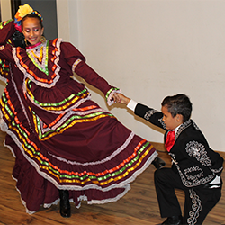 A girl and young boy dance together, both wearing traditional ballet folklorico outfits.