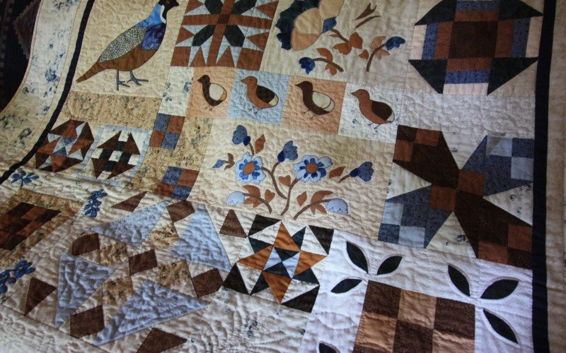 A beige quilt with various squares of bird, plant, and geometric imagery of various blue, brown, and black colors.
