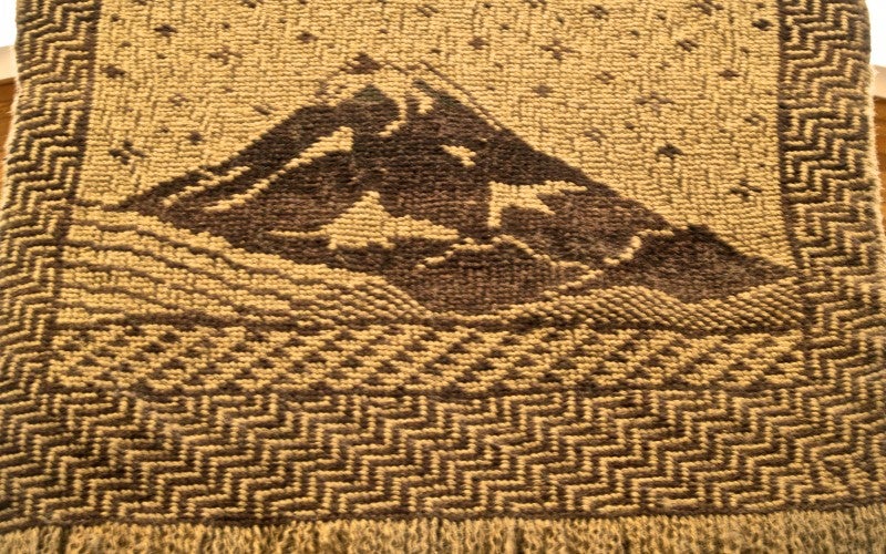 A woven tan tapestry with a purple mountain and a purple zig-zag pattern at the bottom. 