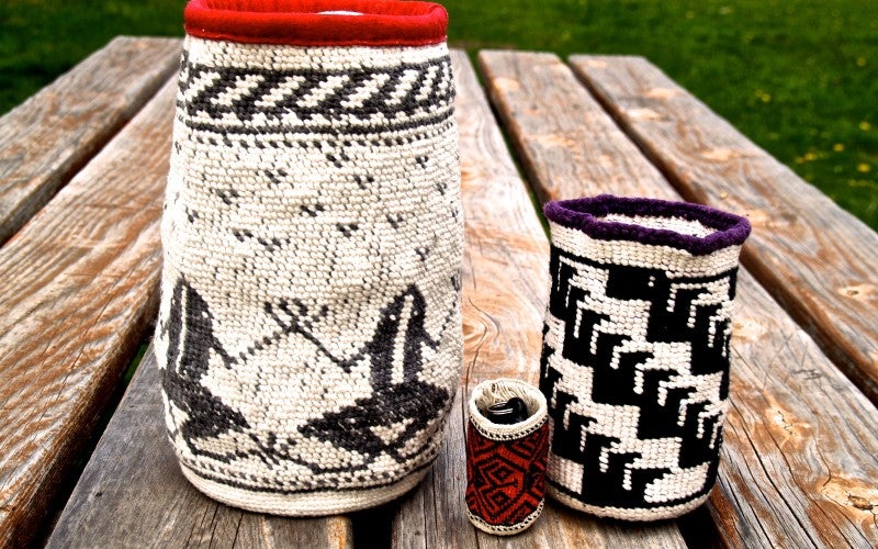 Three woven baskets sit on a wooden table outside. The largest basket is white with red trim and black frogs. The middle sized basket is white with black geometric patterns. The smallest basket is red with white trim and black geometric patterns.