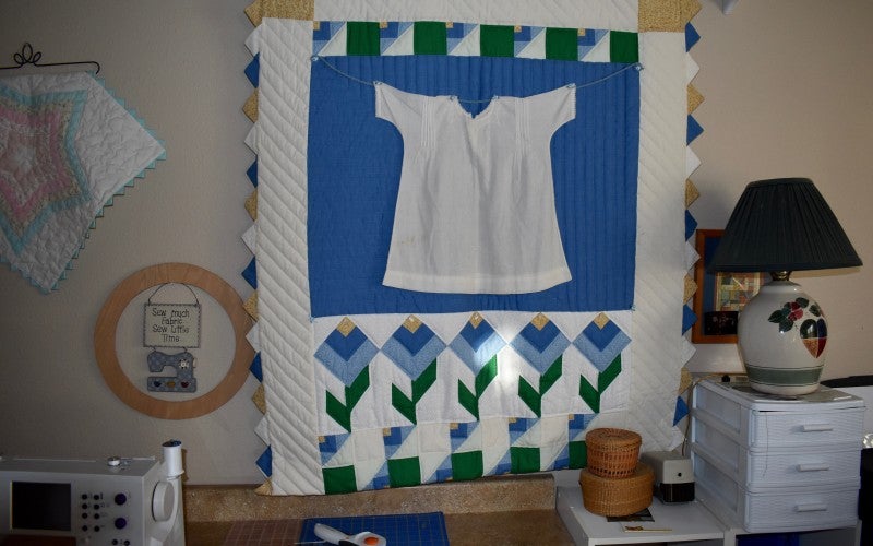 A hanging quilt with a white background, a large blue square in the middle containing a white shirt, and blue, yellow, and green flowers at the bottom.