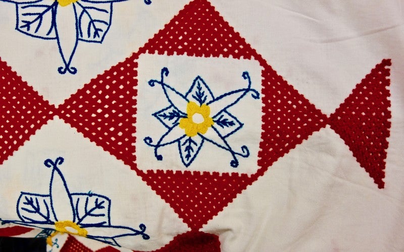 A white sheet with checkered red squares. In the white squares are blue and yellow embroidered flowers.