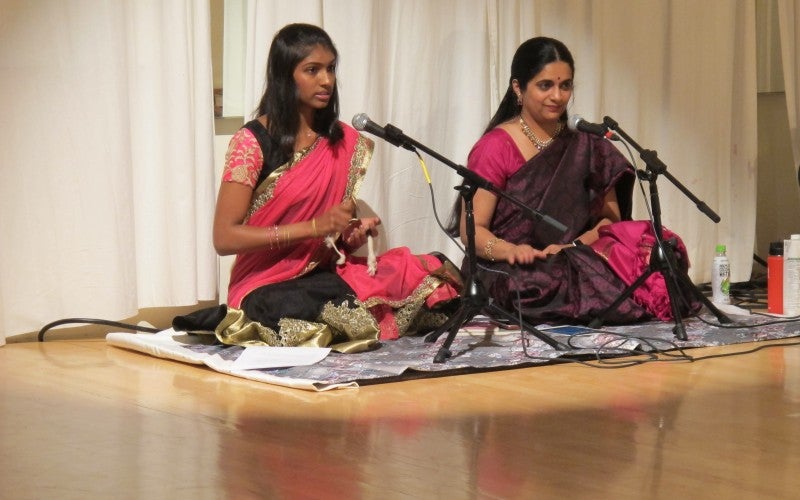 Anita sits in front of a microphone on a dance floor with another girl, both wearing pink traditional outfits.