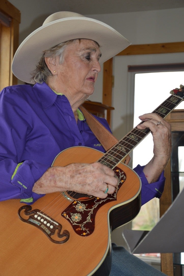 Barbara Nelson sits in a chair playing an acoustic guitar.