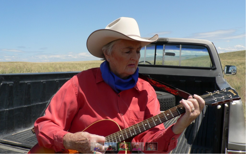 Barbara sits in the back of a truck playing a guitar and wearing a red shirt, blue bandana, and white cowboy hat.