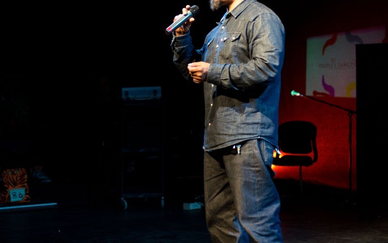 Crenshaw stands on a stage and speaks into a microphone. He wears gray pants, a gray collared shirt, black boots, and a brown baseball cap.