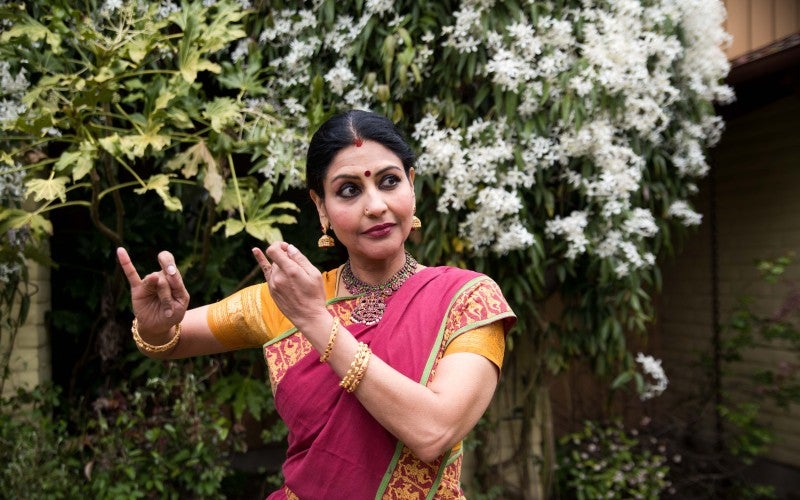 Raman performs a classical Indian dance. She is in front of greenery and wears a yellow and red traditional outfit.