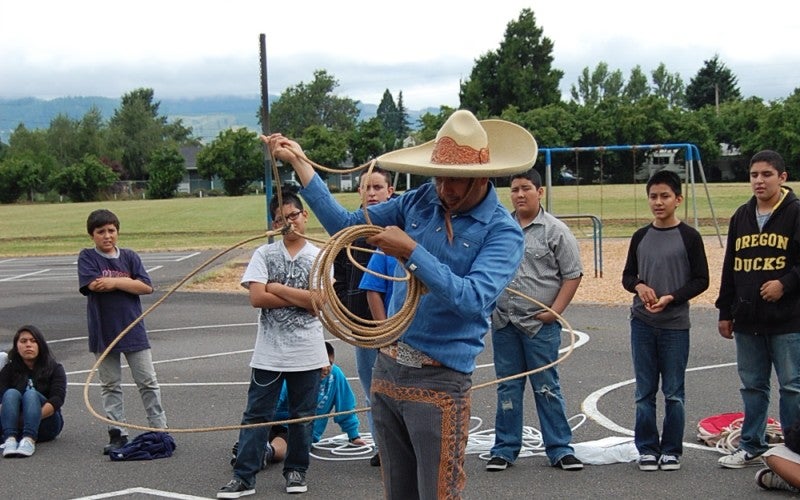 Jose demonstrates cowboy rope work on a playground in front of a group of young boys. He wears a tan cowboy hat, blue button-up shirt, gray pants, and brown boots.