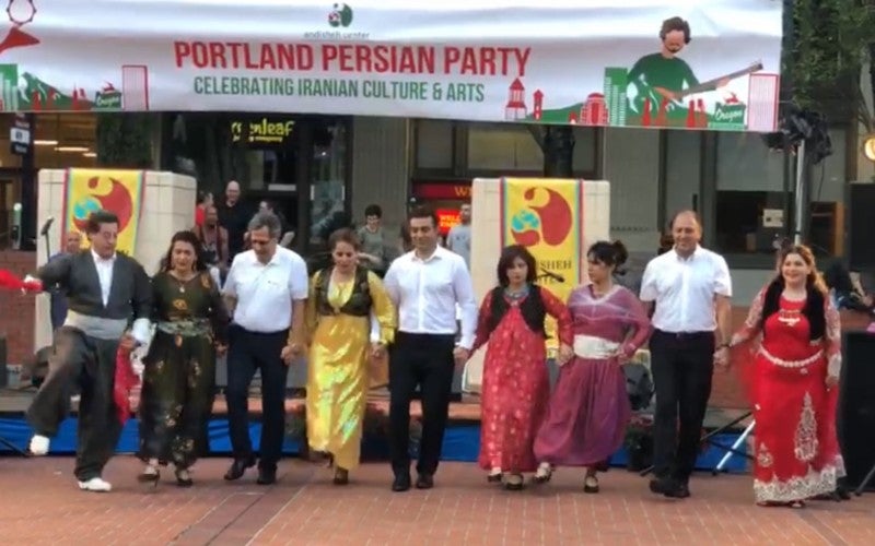 A group of people dressed in regalia line up under white banner that says "Portland Persian Party, celebrating Iranian culture & arts"