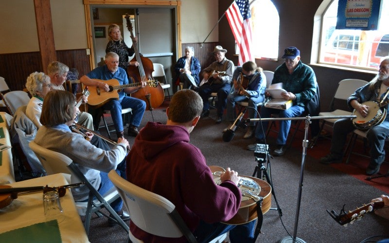Circle of people playing various string instruments inside a room with an American flag hanging on the wall.