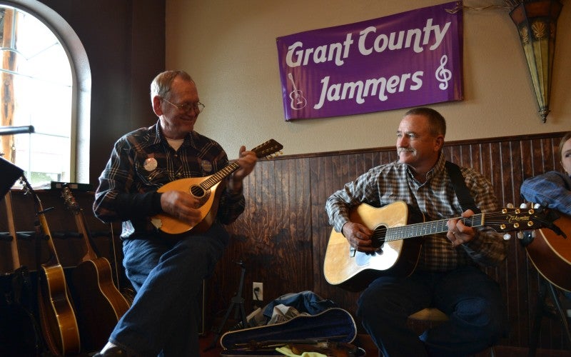 A man playing a guitar accompanied by a man playing a mandolin in a room with a banner reading "Grant County Jammers" on the wall.