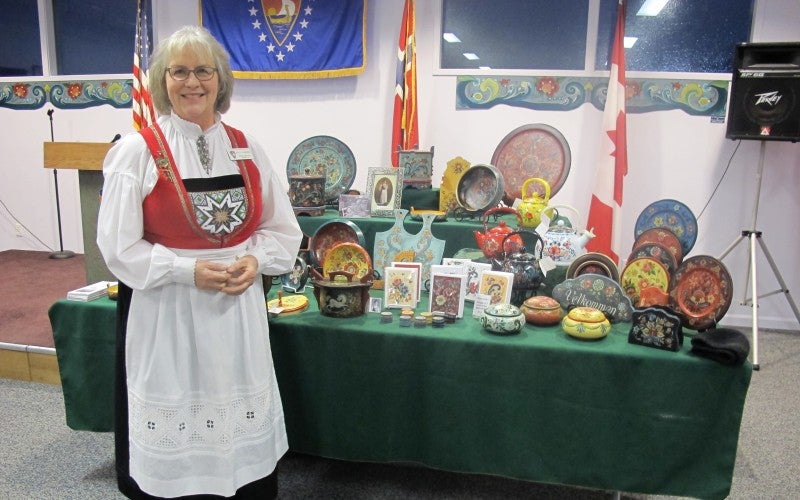 Meshnik stands next to a table displaying her rosemåling work. She wears a traditional Norwegian outfit.