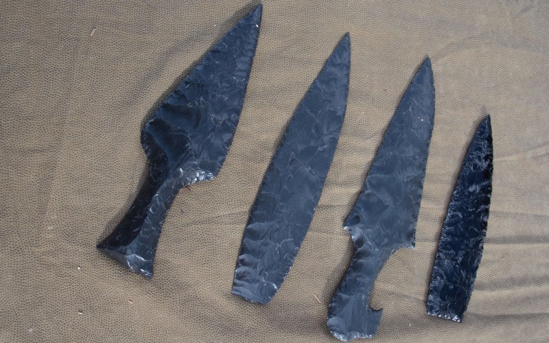 Four larger black obsidian blades sit on a tan cloth.