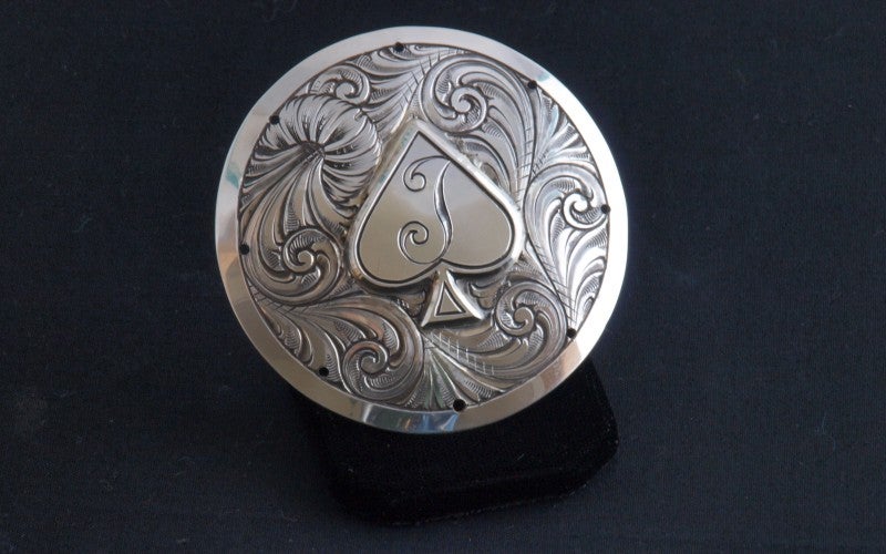 A round piece of engraved silver with a spade in the middle, surrounded by abstract swirls