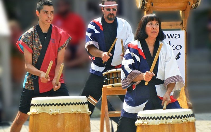 Janet and two other men all play wooden drums while wearing traditional outfits.