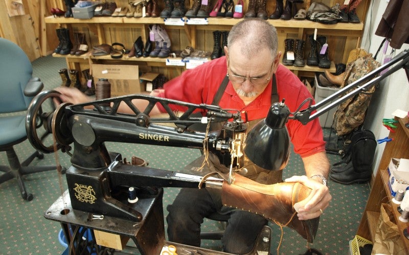 A man sits at a large metal sewing machine and sews a leather cowboy boot together.