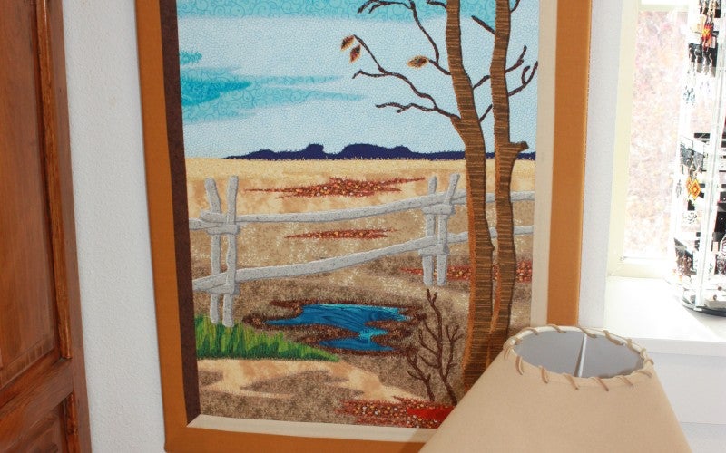 A quilted image of a brown grass landscape. Three birds fly at the top. In the foreground is a leafless brown tree and a white fence with a small puddle next to it.