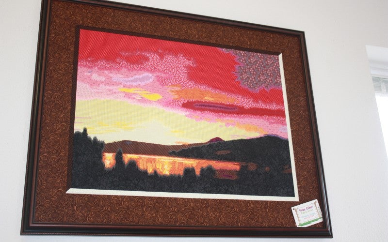 A quilted landscape scene of a lake with a red, pink, and yellow sunset.