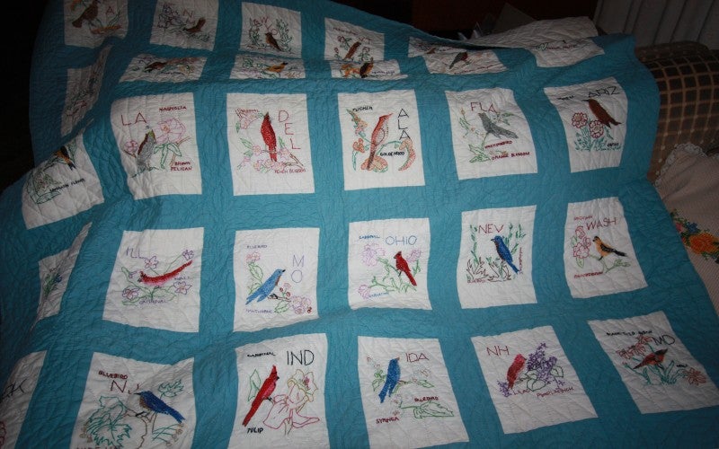 A blue quilt with white squares each containing an embroidered bird.