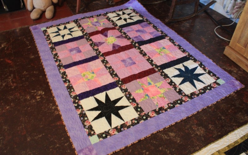 A purple quilt with pink and purple star and cross designs in the middle.