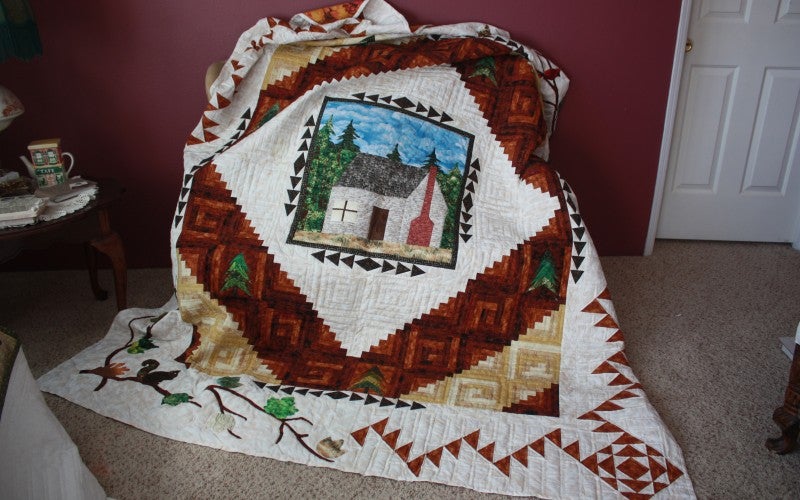 Square white quilt with border decoration and quilted house in the middle.