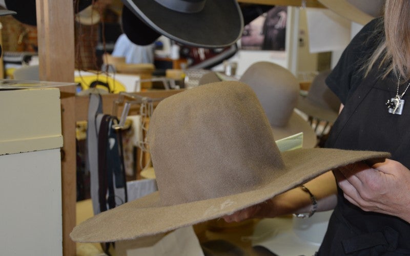 Laura holds a tan cowboy hat and multiple more hats hang on hooks in the background.