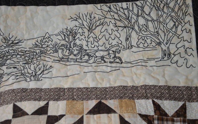 A white quilt containing an embroidered winter scene of a horse drawn sleigh in a forest. 