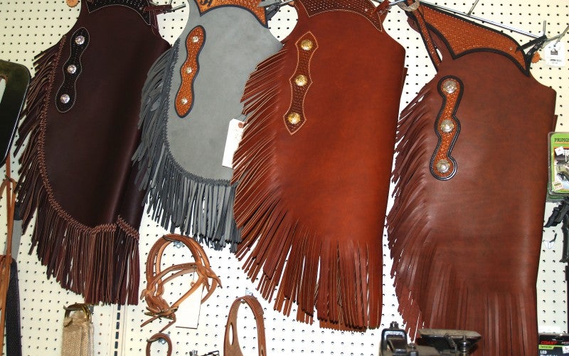Three fringed riding chaps in grey, clay red, and brown.