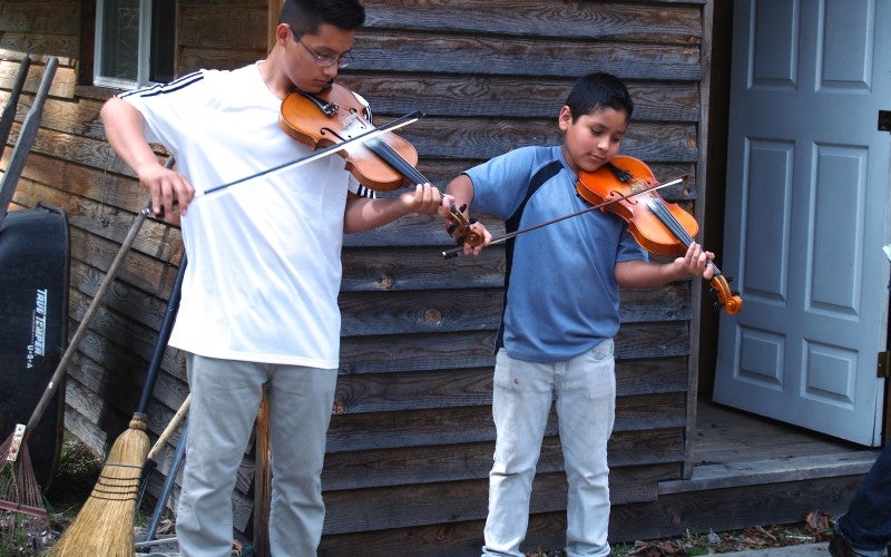 Two boys stand in front of a house playing the violin.
