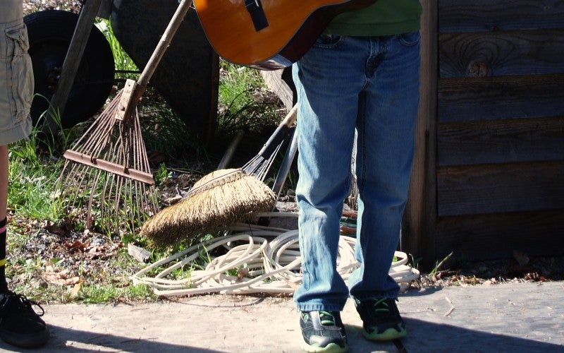 A young boy stands in front of a house and plays a guitar.