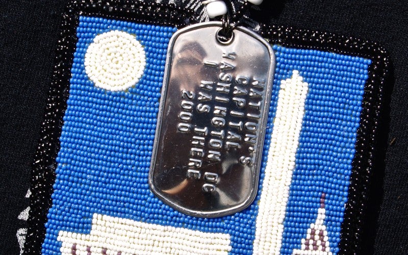 A blue beaded square with white beaded buildings and a silver dog tag on top.