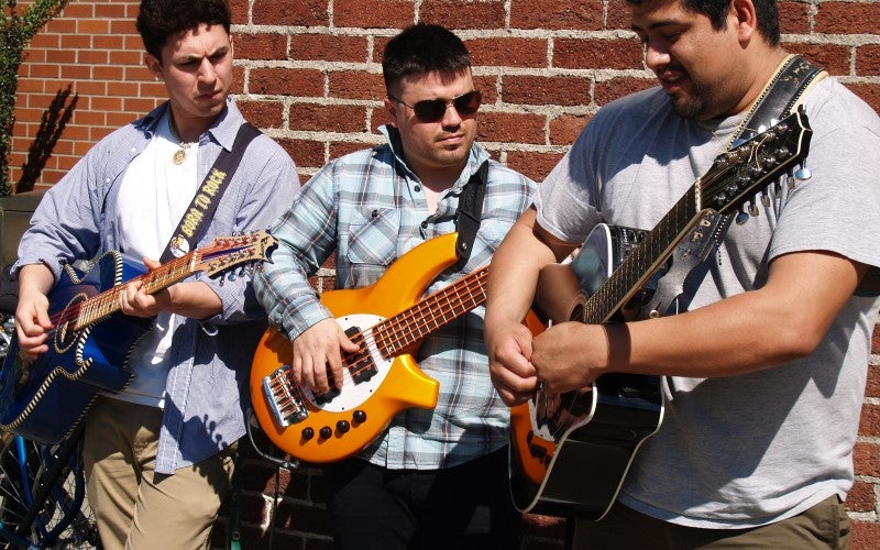 Three men stand in front of a red brick wall and play guitars.