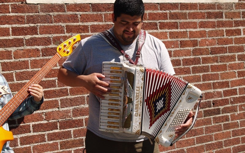A man stands in front of a red brick wall and plays an accordion.