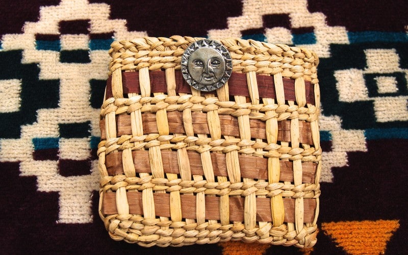 Square shaped woven basket with brown, tan, and red stripes woven into a tan basket with a silver face medallion clasp.