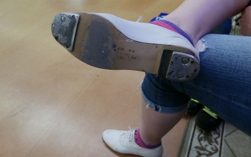 A woman shows the bottom of a white clog shoe, which consists of two metal plates at the toe and heel.