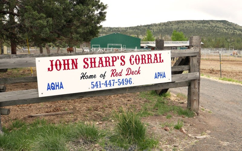 A white sign on a wooden fence saying "John Sharp's Corral."