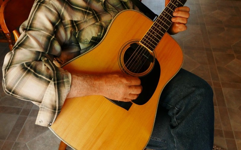Lonny Shurtleff playing a guitar while wearing a white cowboy hat and brown plaid shirt.