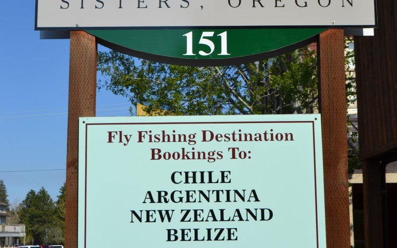 A white shop sign for "The Fly Fisher's Place" located in "Sisters, Oregon." In the middle of the sign is a blue fly fish hook.