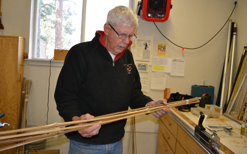 Artist wearing a black jacket and jeans holding three bamboo fishing rods inside his workshop.