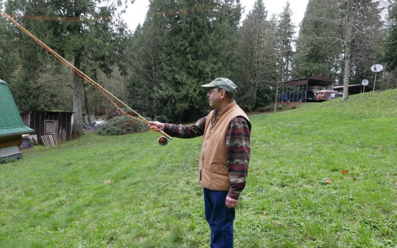 Artist wearing a tan vest and brown plaid shirt posing in a field with a fly-fishing rod.