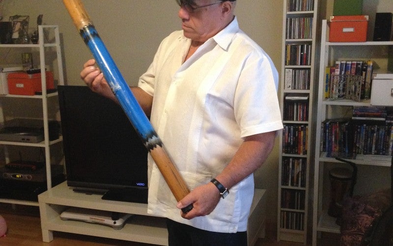 A man stands in a living room wearing a white shirt and black hat. He holds a long, pole-shaped blue instrument.