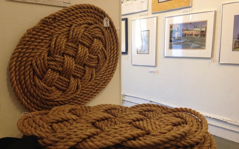 A brown knotted rope mat hangs on a gallery wall, and a pile of more brown knotted rope mats sit on a table in front of the wall.
