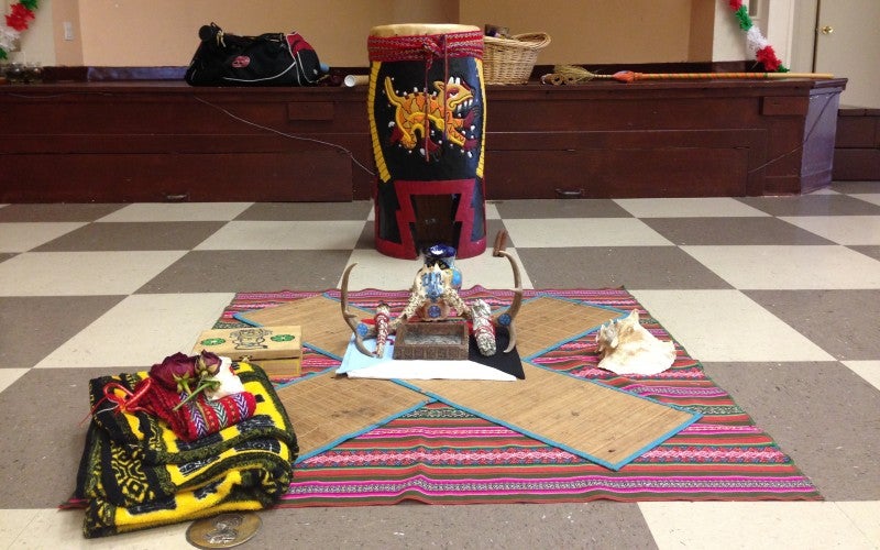 A multi-colored drum sits on a checkered floor. In front of the drum is a pink, red, and green striped bug with various traditional items on top.