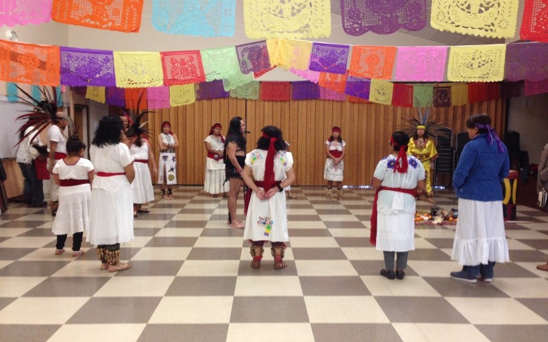 A group of people in a dance room stand in a circle wearing white dresses. The room is decorated with papel picado.